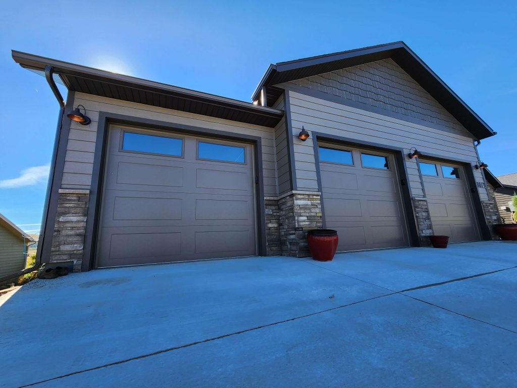 Front view of a three-car garage with tan panel doors, stacked stone accents, barn-style gooseneck wall lights, and red ceramic planters on a wide concrete driveway.
