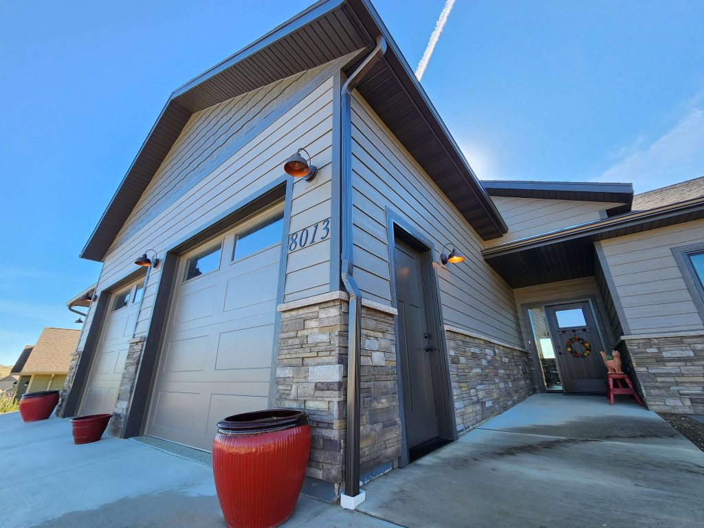 Exterior of a modern home featuring gray lap siding, stacked stone accents, a two-car garage, barn-style wall lights, and a covered front entry with a wreath on the door.