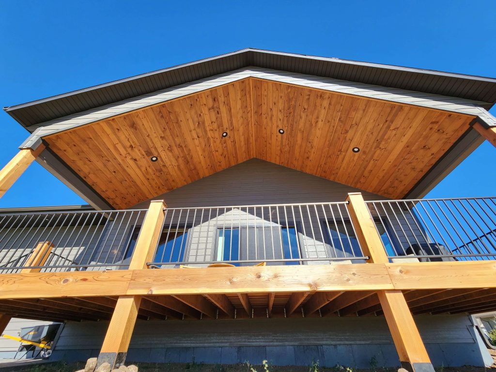 Elevated wood deck with metal railing and a vaulted cedar tongue-and-groove ceiling with recessed lighting, viewed from below against a clear blue sky.