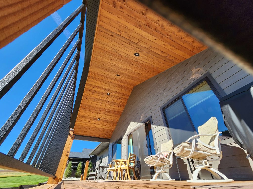 On-deck view of a covered outdoor patio featuring a vaulted cedar tongue-and-groove ceiling with recessed lighting, metal vertical railing, and outdoor furniture including rocking chairs and a wood dining set.