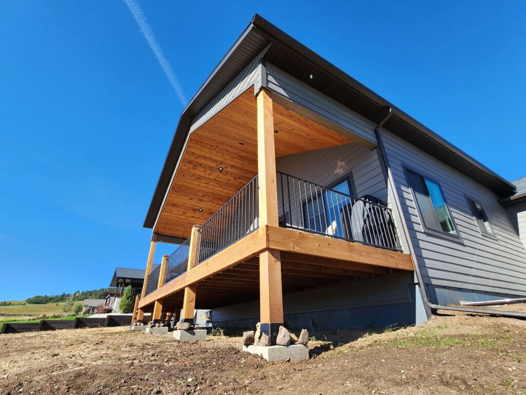 Side-angle view of a home's elevated deck with heavy timber posts, cedar tongue-and-groove covered ceiling, and black metal railing, overlooking a rural landscape under a clear blue sky.